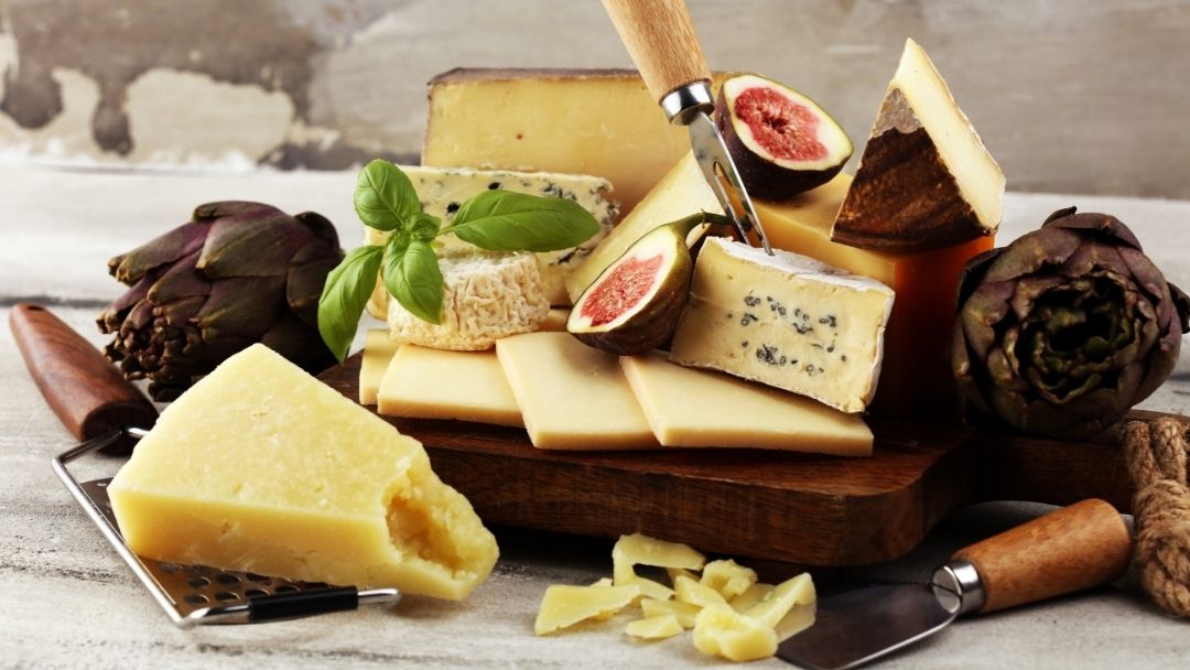 A rustic cheese tasting spread featuring assorted artisan cheeses, figs, basil, and cheese knives arranged on a wooden board, showcasing a variety of textures and flavours for gourmet cheese tasting and entertaining.