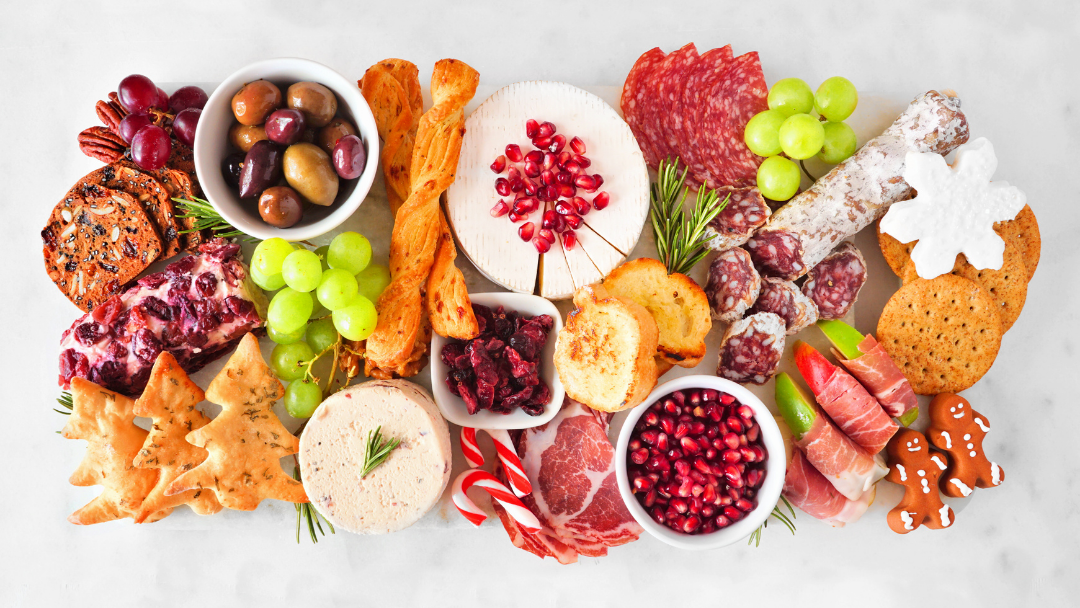 Overhead view of a festive cheese and charcuterie board with soft cheeses, cured meats, olives, crackers, grapes, dried fruit, and seasonal garnishes arranged on a marble surface.