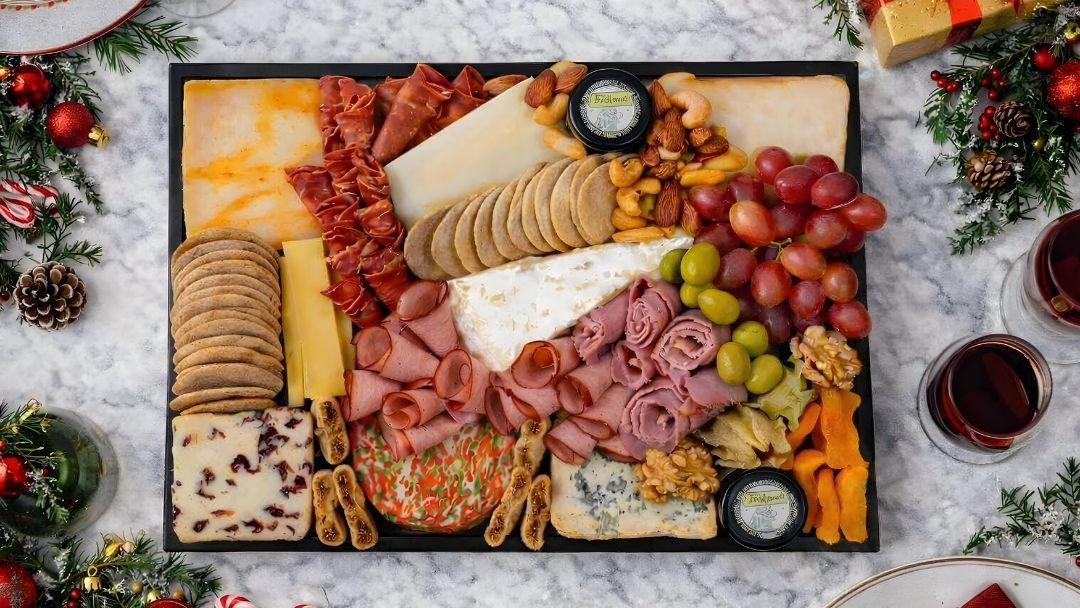 Overhead view of a festive cheese and charcuterie board featuring assorted cheeses, sliced cured meats, crackers, grapes, nuts, and dried fruit on a marble table.