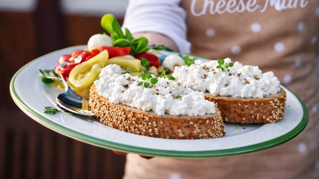 A wholegrain toast topped with fresh cottage cheese, served with sliced vegetables and herbs as a light, healthy cheese meal.