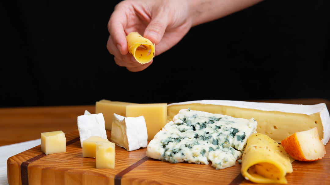 The Cheese Man assorted cheeses by consistency displayed on a wooden board, featuring soft brie, blue cheese, semi-hard cubes, aged wedge, and rolled cheese slices for tasting and pairing.
