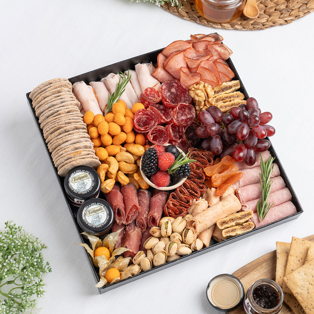 Assorted charcuterie board with meats, cheeses, fruits, and nuts on a white surface. The Cheese Man charcuterie board featuring halal cold cuts, salami, smoked meats, crackers, nuts, dried fruits, grapes, and gourmet accompaniments, curated for luxury grazing platters and cheese boards in Dubai.